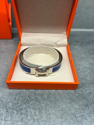 Blått Harmẽs armband i metall - Snyggt armband från Harmẽs i silverfärgad metall med blå emaljdetaljer. Armbandet har den ikoniska H-spännet. Perfekt accessoar för att lyfta din stil. Tyvärr ingen box med då den är borta 