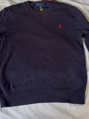 Ralph Lauren tröja  - Mörkblå rundhalsad tröja från Polo Ralph Lauren med röd broderad logga på bröstet. Litet hål vid armhålan.Tröjan har ribbade muddar vid ärmslut och nederkant. Tillverkad i mjukt material som känns skönt mot huden.