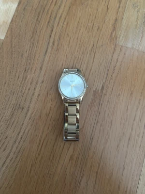 Guldfärgad klocka från Regal - Stilren guldfärgad klocka från Regal med metallarmband och rund urtavla. Urtavlan är vit med gulddetaljer och har en rad små stenar runt kanten för extra bling. Perfekt accessoar för att lyfta din outfit.