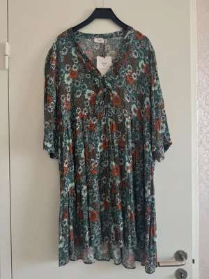 Blommig grön kortklänning - Supersöt kortklänning med grönt och orange blommönster, v-ringning och knytband fram. Klänningen har trekvartsärm och ett luftigt, flowy fall som passar perfekt till varma dagar. Tillverkad i mjuk viskos för skön känsla.