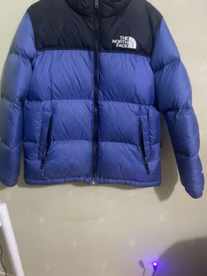 Blå dunjacka från The North Face - Säljer en blå dunjacka från The North Face med svart axelparti och logga på bröstet. Jackan har två dragkedjeförsedda fickor framtill, hög krage och quiltad design. Perfekt för kalla vinterdagar och riktigt snygg streetstil. Perfekt till våren 