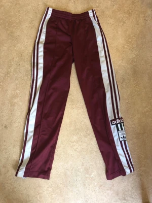 Vinröda Adidas mjukisbyxor med ränder - Säljer ett par vinröda mjukisbyxor från Adidas med klassiska vita ränder längs benen och Adidas-logga på ena benet. Byxorna har resår i midjan och raka ben. Perfekta för en sportig och avslappnad stil.