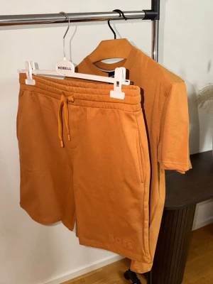 Orange set från BOSS  - Säljer snygg orange set från BOSS med matchande snörning i midjan. Settet har en relaxed passform och är tillverkade i mjuk bomull. Perfekta för sommardagar och har diskret BOSS-logga broderad vid benet.
