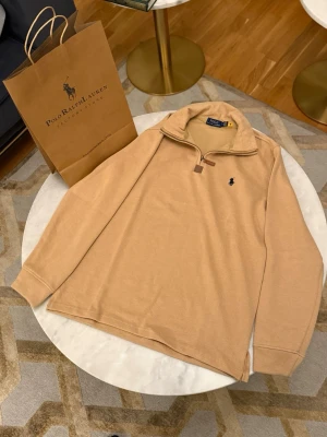 Beige stickad tröja från Polo Ralph Lauren - Snygg beige stickad tröja från Polo Ralph Lauren med halv dragkedja och klassisk broderad logga på bröstet. Tröjan har hög krage och långa ärmar, perfekt för lager på lager. Materialet känns mjukt och bekvämt.