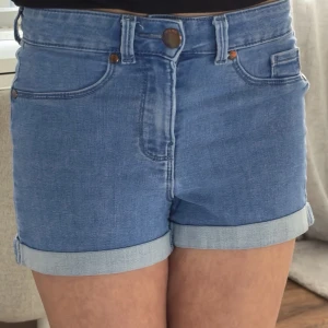 Blå denimshorts med uppvik - Snygga blå denimshorts med uppvikta benslut och klassisk femficksdesign. Shortsen har hög midja, bälteshällor och stängs med knapp och dragkedja. Perfekta för varma dagar och enkla att matcha med olika toppar.