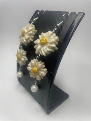 Hängande örhängen med prästkragar - Unika örhängen med dubbla vita prästkragar i plast och gul mitt, avslutade med en liten pärla längst ner. Blommorna har en skimrande yta och örhängena har krokar i metall. Perfekta för dig som vill sticka ut med ett lekfullt och somrigt smycke.