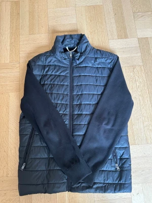 Svart quiltad cardigan med dragkedja - Snygg svart cardiganjacka med quiltad framsida och stickade ärmar samt rygg. Jackan har hög krage, två fickor med dragkedja och hel dragkedja framtill. Perfekt för lager-på-lager och en clean look. Stolek XL men passar L.