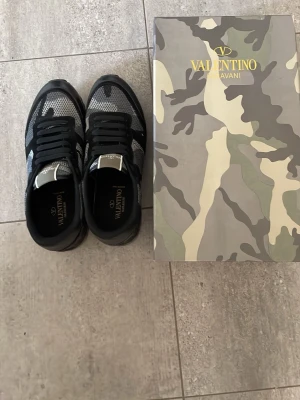 Valentino Garavani svarta sneakers - Snygga svarta sneakers från Valentino Garavani med grått och svart camouflagemönster. Skorna har meshpaneler, mockadetaljer och svarta snören. Yttersulan är räfflad för extra grepp och designen är sportig men exklusiv.