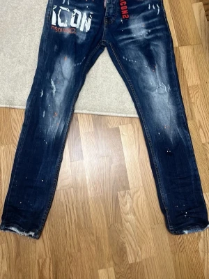 Mörkblå Dsquared2 ICON jeans - Säljer ett par mörkblå Dsquared2 ICON jeans med skinny fit och slitna detaljer. Jeansen har vit och röd text framtill, målade stänk och fransade benslut. Klassisk femficksmodell i denim med cool streetwear-vibe.