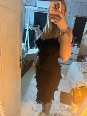 Svart volangklänning med smala axelband - Säljer en svart klänning med smala axelband och volangdetaljer längs hela klänningen. Modellen är figurnära och har en feminin siluett som slutar strax under knät. Perfekt för dig som gillar en elegant och trendig look.