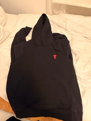 Svart hoodie från Ami Paris med röd logga - Svart hoodie från Ami Paris med klassisk huva och stor magficka. På bröstet sitter Ami:s ikoniska röda hjärta med A. Tillverkad i mjukt bomullsmaterial som känns skönt mot huden. Perfekt för en avslappnad och trendig look.