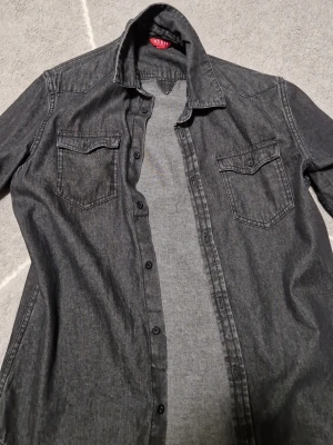 Svart jeansjacka från Guess XS - Snygg svart jeansjacka från Guess i storlek XS. Klassisk modell med två bröstfickor med lock och knappar framtill. Jackan har en avslappnad passform och är tillverkad i slitstarkt jeansmaterial. Perfekt att slänga över en t-shirt för en cool look.