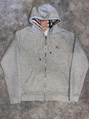 Grå zip hoodie från Burberry - Snygg grå zip hoodie från Burberry med klassiskt rutigt foder i huvan och broderad logga på bröstet. Tillverkad i mjuk bomullsblandning, har dragsko i huvan och två stora fickor framtill. Perfekt för en chill och stilren look.