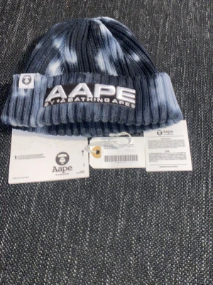 Blå tie-dye mössa från AAPE - Snygg ribbstickad mössa från AAPE by A Bathing Ape i blått och vitt tie-dye-mönster. Stor logotyp framtill och liten patch på sidan. Tillverkad i mjukt material som håller dig varm och ger en streetwear-vibe.