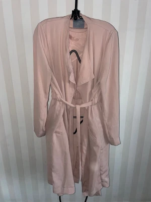 Ljusrosa trenchcoat med skärp storlek S-M - Stilren ljusrosa trenchcoat från Lindex med avslappnad passform, bred krage och knytskärp i midjan. Jackan har två fickor framtill och är tillverkad i ett mjukt, tunt tyg som ger ett snyggt fall. Perfekt för vår och höst.