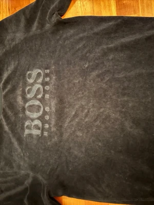 Svart sweatshirt från Hugo Boss - Snygg svart sweatshirt från Hugo Boss med stor BOSS-logga broderad på bröstet. Tröjan har rund halsringning, långa ärmar och ribbade muddar vid ärmslut och nederkant. Mjuk och skön kvalitet, perfekt för en avslappnad stil. Väldigt najs material.