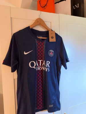 PSG matchtröja Nike blå röd - Snygg PSG matchtröja i mörkblått med röd och vit detalj på bröstet. Tröjan har klubbmärke, Nike-logga och Qatar Airways-tryck framtill. Tillverkad i lätt och ventilerande material, perfekt för fotboll eller träning.