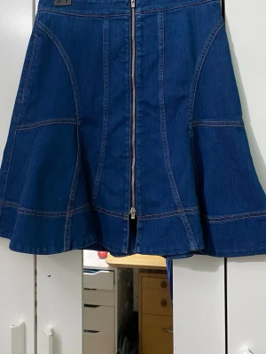 Blå jeanskjol med dragkedja - Säljer en snygg blå jeanskjol med markerade sömmar och hel dragkedja framtill. Kjolen har en utställd form som ger en cool siluett och passar perfekt till en avslappnad stil. Perfekt att matcha med sneakers eller boots. Midjan är 35cm och längden är 51cm. 