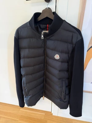 Svart cardigan från Moncler - Snygg svart cardigan, från Moncler med quiltad front och ärmar i mjukare material. Jackan har dragkedja framtill, ståkrage och Moncler-logga på bröstet. Perfekt för kalla dagar och ger en clean look. Storlek 4 vilket är det samma som large. Passar både large och medium.