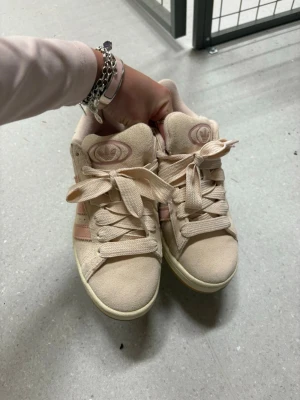 Beige Adidas Campus sneakers - Snygga Adidas Campus sneakers i beige mocka med ljusrosa detaljer och klassiska tre ränder på sidan. Skorna har en låg profil och gummisula, samt Adidas-logga på plösen. Perfekta för en chill och trendig stil.