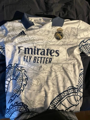 Real Madrid fotbollströja #10 Modric - Säljer en vit Real Madrid matchtröja från Adidas med blått drakmönster, klubbmärke och sponsortryck framtill. Krage i blått och namn samt nummer 10 'Modric' på ryggen. Perfekt för dig som älskar fotboll och vill sticka ut på planen.