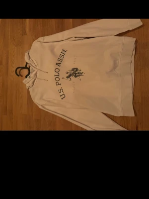 Vit hoodie från U.S. Polo Assn. - Snygg vit hoodie från U.S. Polo Assn. med tryck framtill och klassisk huva med dragsko. Hoodien har ett stort logotryck med hästmotiv och texten 'U.S. POLO ASSN.' på bröstet. Mjuk insida och ribbade muddar vid ärmslut och nederkant.