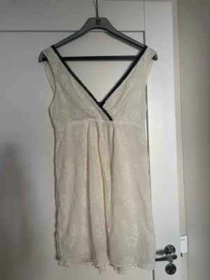 Krämvit klänning med spetsdetalj - Klänning i krämvitt med broderat blommönster och svart spets längs den djupa v-ringningen. Ärmlös modell i lätt, tunt tyg som ger en romantisk känsla. Perfekt för mysiga kvällar hemma.