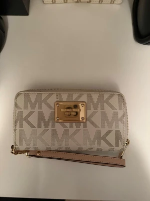 Beige plånbok Michael Kors med logga - Snygg beige plånbok från Michael Kors med guldiga detaljer och stor MK-logga över hela ytan. Plånboken har dragkedja runtom, flera fack för kort och sedlar samt en handledsrem. Tillverkad i tåligt konstläder med stilren design.