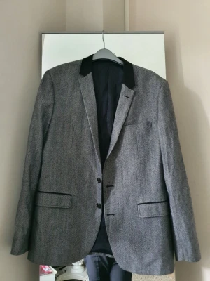 Grå kavaj från Matinique - Stilren grå kavaj från Matinique med svart krage och två knappar framtill. Kavajen har två fickor med lock och en bröstficka. Insidan är fodrad med randigt tyg och har en innerficka. Perfekt för en dressad look.