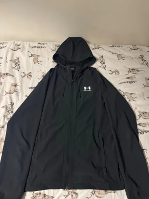 Svart vindjacka från Under Armour - Snygg svart vindjacka från Under Armour med dragkedja framtill och justerbar huva. Jackan har vit logga på bröstet och meshfoder i huvan för extra komfort. Perfekt för blåsiga dagar och sportiga outfits. Har även kvitto 
