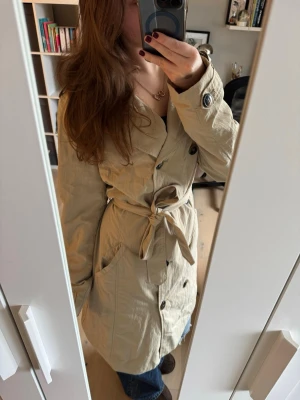 Beige trenchcoat från Kappahl - Stilren beige trenchcoat från Kappahl med dubbelknäppning, knytskärp i midjan och klassisk krage. Jackan har svarta knappar, axelklaffar och stora fickor framtill. Perfekt för dig som vill ha en tidlös och trendig look.