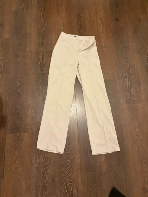 Vida beige kostymbyxor - Snygga beige kostymbyxor med hög midja och vida ben. Byxorna har pressveck framtill och stängs med knapp och dragkedja. Perfekta för en clean och stilren look.