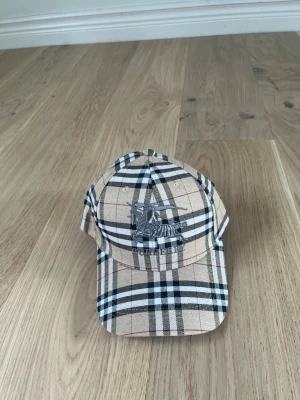 Beige Burberry keps med rutmönster - Säljer en klassisk beige keps från Burberry med det ikoniska rutmönstret i svart, vitt och rött. Kepsen har broderad logga framtill och justerbar rem bak. Tillverkad i bomull och perfekt för dig som vill ha en stilren accessoar med lyxig känsla.