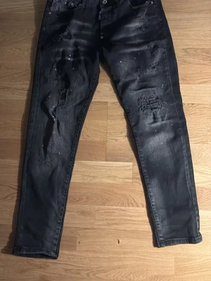 Svarta disquared - Säljer ett par svarta skinny jeans med slitningar och hål på benen samt små vita färgstänk. Jeansen har fem fickor och knappgylf. Snygg tvättad look och smal passform som ger en trendig vibe har även hål i fickan där man inte kan lägga in sin mobil bara i den ena fickan..