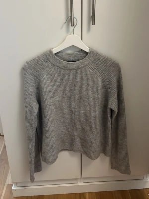 Grå stickad tröja från Pieces - Mysig grå stickad tröja från Pieces med rund halsringning och ribbstickad struktur. Tröjan har långa ärmar och en avslappnad passform, perfekt för kyliga dagar. Enkel och stilren design som passar till det mesta. Har en liten fläck vid halsen