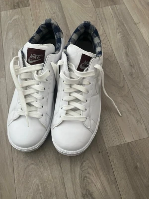 Vita Nike sneakers med rutigt foder - Snygga vita Nike sneakers i läder med klassisk låg profil och vita snören. Insidan har ett rutigt foder i blått och vitt som ger en unik touch. Märkeslogga på plösen i vinrött. Perfekta för dig som vill ha en clean och stilren look.