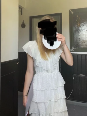 Vit volangklänning med prickigt mönster - Söt vit kortklänning med volanger och diskret prickigt mönster. Klänningen har v-ringning, ärmlös design med breda volangärmar och flera lager volanger i kjolen. Perfekt för sommarens tillfällen och studentfirande.