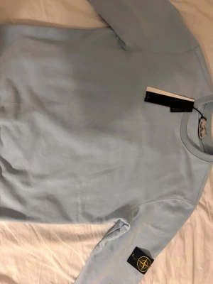 Ljusblå sweatshirt från Stone Island, priset kan diskuteras - Snygg ljusblå sweatshirt från Stone Island med klassisk logotyp-patch på ärmen. Tröjan har rund halsringning och raka ärmar. Perfekt för dig som gillar streetwear och vill ha en stilren look.