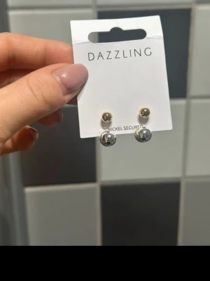 Dubbla studsörhängen från Dazzling - Säljer ett par snygga dubbla studsörhängen från Dazzling. Örhängena har en liten guldfärgad kula framtill och en större silvrig kula baktill. Klassisk och stilren design som passar till många looks.