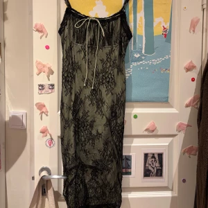 Lång svart spetsklänning med knyt - Långklänning med svart spets över ljust tyg, tunna axelband och dekorativt knytband framtill. Klänningen är ärmlös och har en romantisk vibe med transparent spets och lätt material. Perfekt för dig som gillar detaljer.