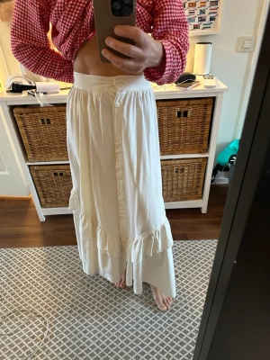 Vit långkjol med volang och knappar Stl m - Säljer en creme vit långkjol med knappar framtill och volangdetaljer längs nederkanten. Kjolen har en avslappnad passform och är tillverkad i ett mjukt bomullstyg. 100 % bomull