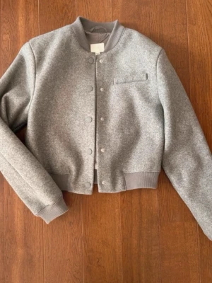 H&M grå bomberjacka - Stilren grå bomberjacka med ribbade muddar och krage. Jackan har knappar framtill och en minimalistisk bröstficka. Perfekt för dig som gillar en enkel men trendig look. Aldrig använd! Säljes/ strl S