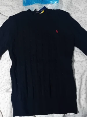 Svart kabelstickad tröja Polo - Kabelstickad tröja i mörkblått från Polo  med klassisk rund halsringning och den ikoniska röda logotypen broderad på bröstet. Tröjan har långa ärmar och ribbade muddar för en snygg och bekväm passform.