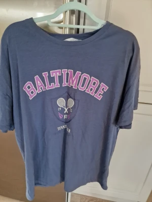 Blå t-shirt Baltimore Primark - Blå t-shirt från Primark med trycket 'BALTIMORE' i rosa och vitt samt tennismotiv framtill. Skön och avslappnad passform, perfekt för en sportig look. Tillverkad i mjuk bomull och har klassisk rund hals och korta ärmar. I mycket gott skick 