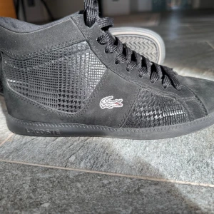 Svarta höga sneakers från Lacoste - Snygga svarta höga sneakers från Lacoste med klassisk krokodillogga på sidan. Skorna har en mix av mocka och syntetmaterial med svagt mönster ton-i-ton på sidorna, rund tå och platt sula. Perfekta för hösten!