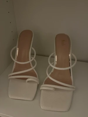 Vita trendiga pumps med remmar - Säljer ett par vita pumps med smala, rundade remmar och fyrkantig tå. Skorna har öppen häl och hög klack, vilket ger en modern och stilren look. Perfekta för dig som vill sticka ut med en minimalistisk och chic stil. Materialet ser ut att vara konstläder.