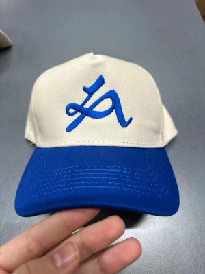 Beige och blå keps med broderad logga - Snygg keps i beige med blått broderat motiv framtill och blå skärm. Justerbar snapback baktill för perfekt passform. Tillverkad i bomull, perfekt för dig som vill ha en stilren och sportig look.