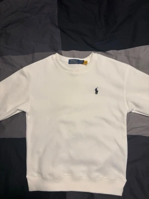 Vit sweatshirt från Polo Ralph Lauren - Klassisk vit sweatshirt från Polo Ralph Lauren med rund halsringning och broderad logga på bröstet. Tröjan har ribbade muddar vid ärmslut och nederkant, perfekt för en clean och stilren look.