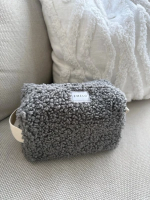 Grå fluffig necessär från Emelo - Supermjuk och fluffig necessär i grå teddy från Emelo. Praktisk rektangulär form med vit dragkedja och ett brett, beige handtag på sidan. Perfekt för att förvara smink eller småprylar. Stilren och mysig design som sticker ut. Storlek medium. Mått ca 20x12x8cm.