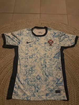 Portugal Nike fotbollströja blåvit - Säljer en Portugal fotbollströja från Nike i blått och vitt med grafiskt mönster. Tröjan har svarta detaljer vid ärmslut och sidor samt Portugal-emblem på bröstet. Perfekt för dig som älskar fotboll och vill sticka ut på planen.